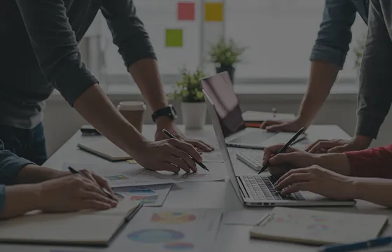 Primer plano de personas trabajando en un proyecto de marketing, mostrando las manos de cuatro personas con bolígrafos apuntando sobre papel y computadoras portátiles.